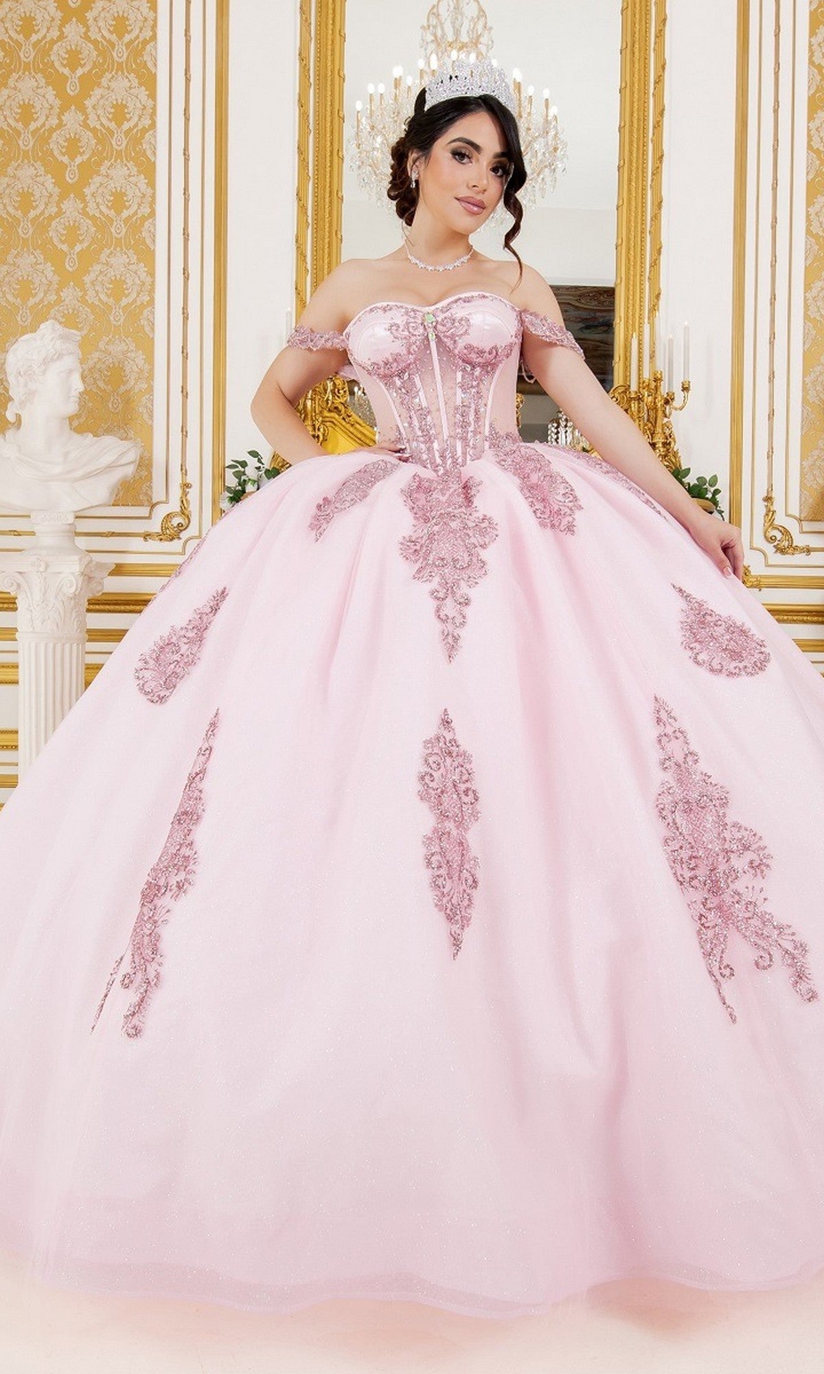 A young woman in a pink Quinceanera Dress 8190J by Cinderella Couture, adorned with floral details, stands in an ornate room featuring gold accents, a chandelier, and a white statue in the background. The elegant off-the-shoulder ball gown adds a fairytale touch to the enchanting scene.